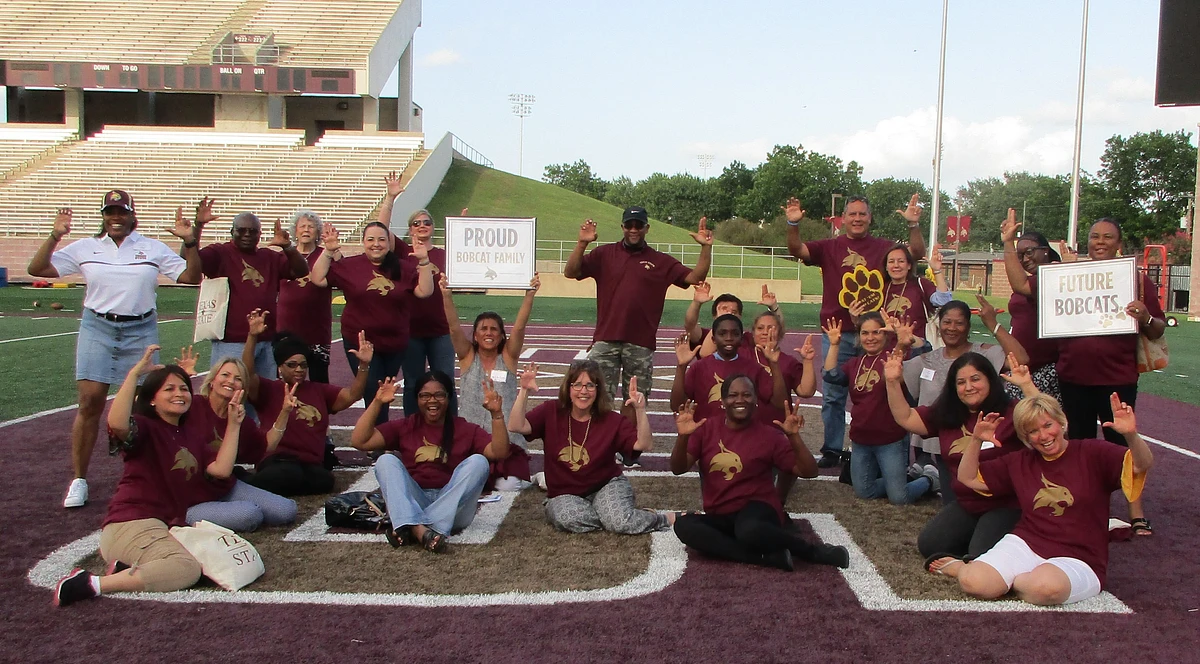 Group of families sitting and standing on the football field shoiwng their TXST hand signs