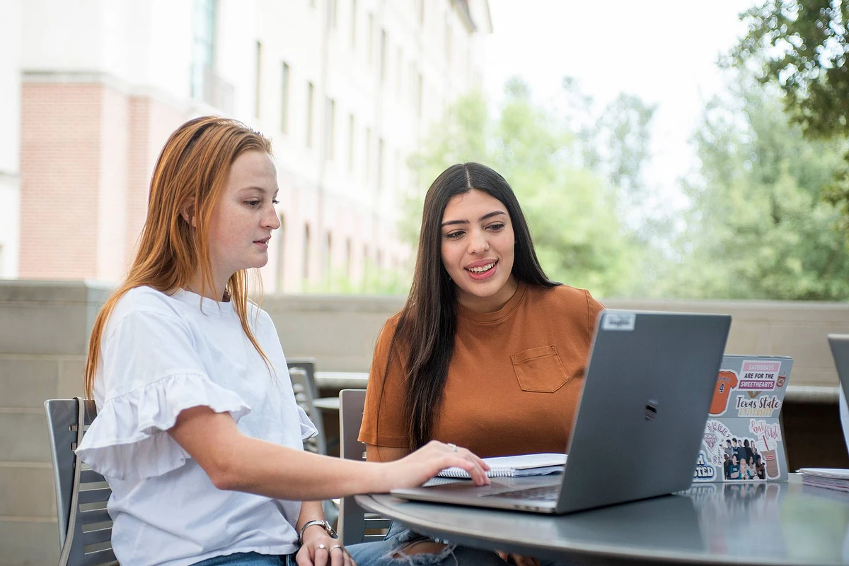 Two women are sitting outdoors, working on laptops and appearing to discuss something.