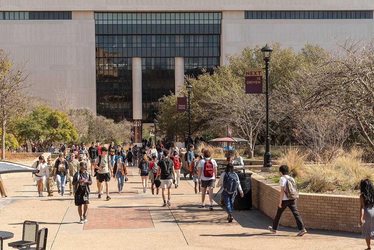 A group of people walks along a campus pathway toward a large building, surrounded by trees and banners.