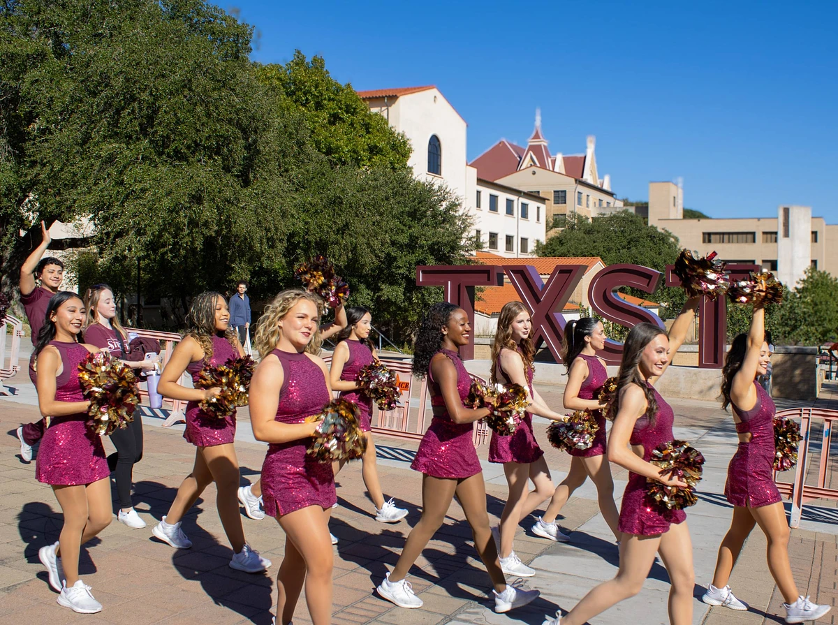 A group of cheerleaders in maroon outfits walk energetically past large "TXST" letters on a sunny day.