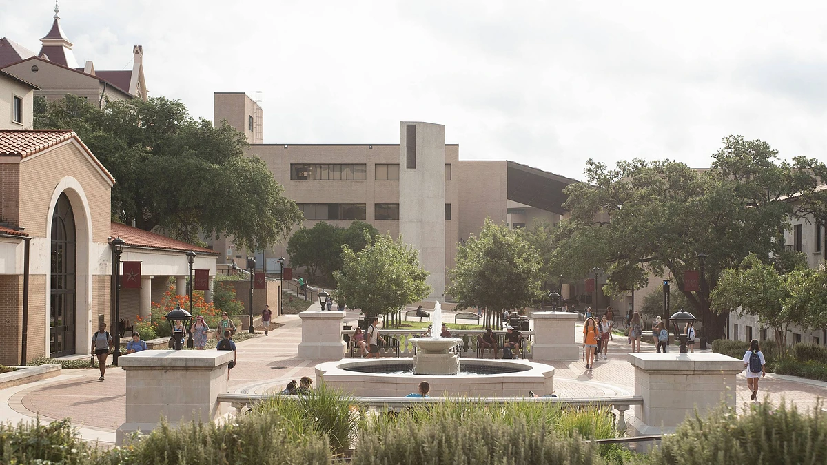 The image shows a bustling outdoor area of a campus with students walking around a central fountain, surrounded by trees and buildings.