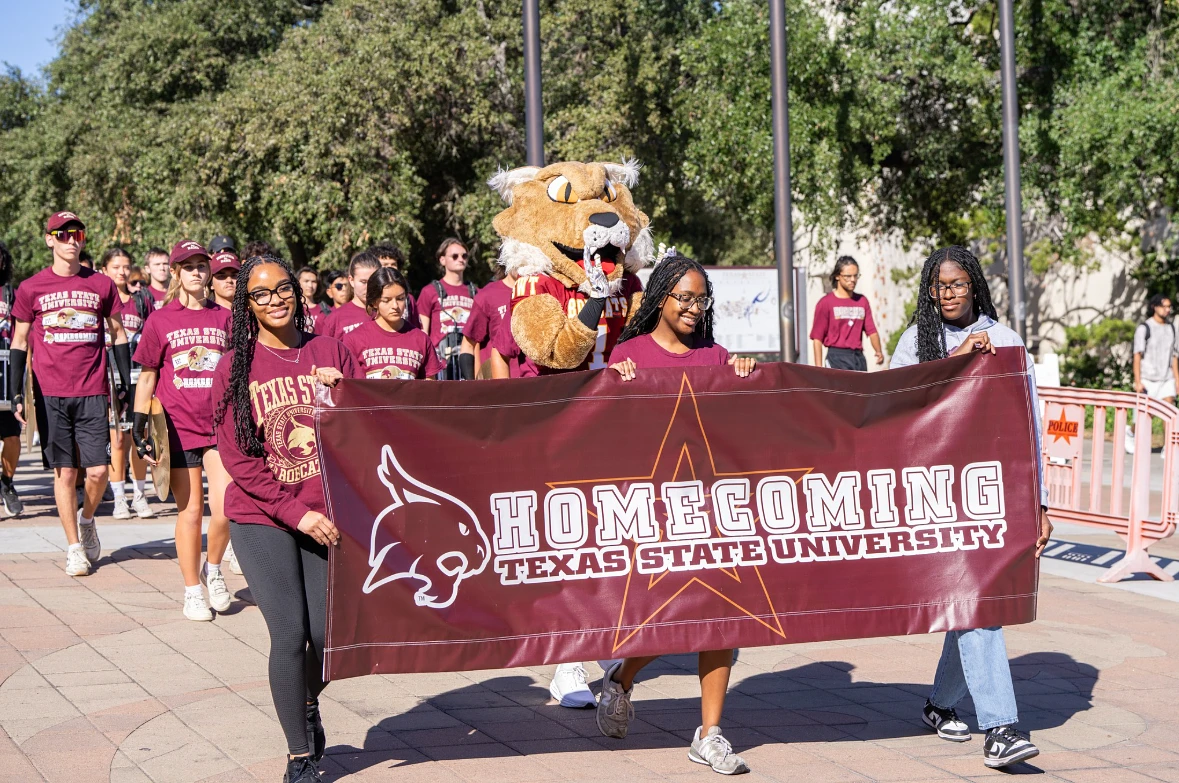 A group of students and a mascot are participating in a parade while holding a "Homecoming Texas State University" banner.